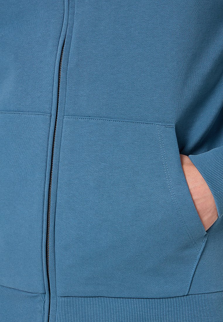 Niebieska bluza z zamkiem błyskawicznym, z ściągaczami na mankietach i dolnym obszyciu, pokazująca rękę częściowo włożoną w kieszeń z przodu po prawej stronie.