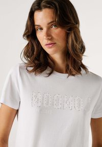 Balta kokvilnas t-kreklu ar rhinestone rotājumu "LONDON" druku, apaļu kakla izgriezumu un īsām piedurknēm. Gluda tekstūra, ikdienišķa piegriezne.