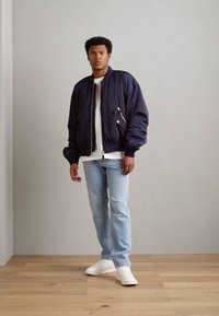 Chaqueta de bombardero navy con corte oversize, con bolsillos de cremallera y botón. Combinada con jeans de color azul claro y zapatillas blancas sobre un suelo de madera clara.