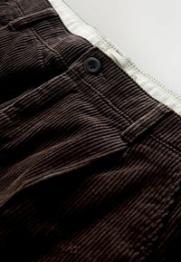Close-up of dark brown corduroy pants waistband with button and beige inner lining on a white background.