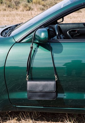 Un sac à bandoulière noir est accroché au rétroviseur latéral d’un cabriolet bleu sarcelle garé dans un champ herbeux.