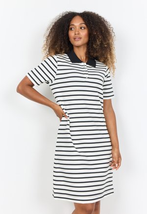 Vrouw met krullend haar die een wit-zwarte horizontaal gestreepte polo jurk met een zwarte kraag draagt, staand met één hand op haar heup.