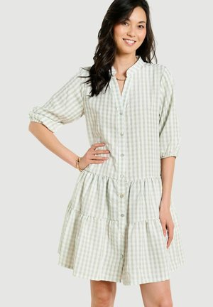 Kobieta uśmiechnięta, ubrana w jasnorozkrojoną, zielono-białą sukienkę w kratę z guzikami, z warstwową spódnicą i rękawami do łokcia, pozująca z jedną ręką na biodrze.