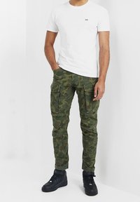 Zelené maskáčové kalhoty s kapsami, doplněné bílým tričkem s kulatým výstřihem. Celý outfit zakončují černé tenisky.