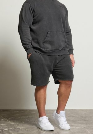 Mężczyzna ubrany w węglonoszary sweter z kieszenią z przodu, pasujące szorty i białe trampki stoi na szarej podłodze na tle jednolitej ściany.