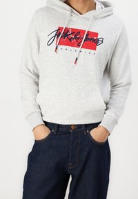 Ljusgrå hoodie med en framficka, dragsnören och en röd och marinblå logotyp. Buren med mörk denimjeans som visar blått tygmönster och sömnad.