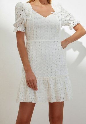 Vrouw draagt een witte jurk van eyelet-stof met opgeblazen korte mouwen, een gerimpelde zoom en een sweetheart-hals met strikdetails.
