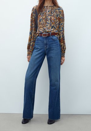 Femme portant un jean bleu taille haute à jambes larges, ceinture marron, blouse à manches longues à motif floral, et chaussures pointues foncées.