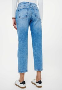 Kvinna som bär hög midja, ljusblå cropped jeans och vita sneakers, stående mot en enkel vit bakgrund, sett bakifrån.