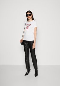 T-shirt bianco in cotone con un logo triangolare rosso con il testo "GUESS?", abbinato a pantaloni in pelle nera e stivaletti neri a punta.