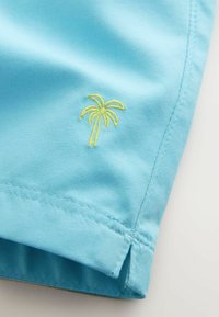 Tissu bleu clair avec ourlet cousu et petite broderie de palmier jaune-vert près du bord.