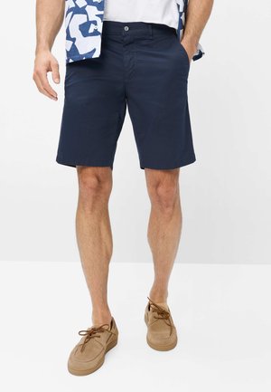 Mann trägt marineblaue Shorts, braune Schnürschuhe, weißes T-Shirt und ein blau gemustertes offenes Hemd mit Händen in den Taschen.