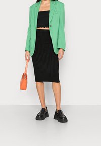 Blazer verde de gran tamaño, top corto de canalé negro, falda midi de canalé negro, bolso de textura naranja y zapatos de plataforma negros.