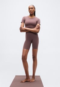 Καφέ κορμάκι με κοντά μανίκια, εφαρμοστή σχεδίαση και λεία υφή, σετ με ταιριαστά σορτς, φορεμένο από μοντέλο που στέκεται σε καφέ επιφάνεια.