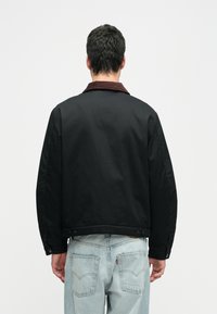 Vyras, dėvintis juodą striukę su rudais apykaklėmis ir šviesiai mėlynomis džinsinėmis kelnėmis, matomas iš nugaros priešais paprastą baltą foną.