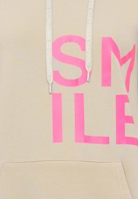 Beige hooded sweatshirt with large pink "SMILE" print. Features white drawstrings with "HAPPINESS" text and a front pocket. Soft material.