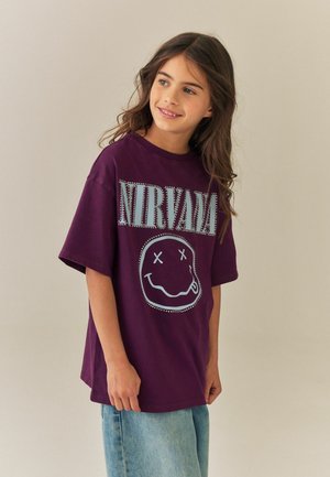 Smilende ung pige med langt brunt hår iført en lilla Nirvana t-shirt og lyseblå jeans, stående mod en ensfarvet baggrund.