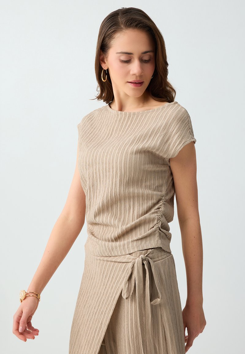 Femme portant un haut beige texturé à manches courtes et une jupe assortie avec un lien sur le côté, regardant vers le bas sur un fond gris clair uni.