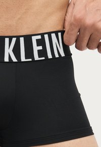 Nærbillede af en person, der justerer sorte boxershorts med en bred linning med fed hvid tekst "KLEIN".