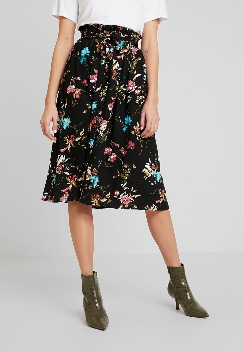 Zwarte bloemenrok met een geplooide tailleband en strikdetail, met levendige roze en blauwe bloemen op een soepele stof. Een paar olijfgroene enkellaarsjes zichtbaar.