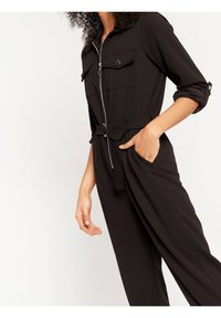 Black jumpsuit with a zip front, two chest pockets, rolled sleeves, and a belted waist. Made from smooth fabric, featuring a relaxed fit.