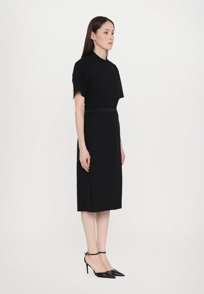 Schwarzes knielanges Kleid mit kurzen Ärmeln und tailliertem Schnitt. Besitzt eine elegante Silhouette und subtile seitliche Schlitze. Getragen mit spitzen schwarzen Absätzen.