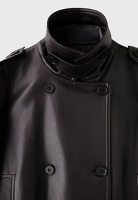 Manteau en cuir noir à double boutonnage avec col montant, pattes d’épaule boutonnées et gros boutons noirs à l’avant et au col.