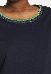 Námořnická košile s kulatým výstřihem, která obsahuje zelený a metalický akcent. Měkčená textilie, viditelné stehy podél límce.