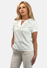 Biała bluzka z krótkim rękawem o dekolcie w kształcie litery V i plisowanymi detalami. Gładka tkanina i luźny krój. Zestawiona z jasnobeżowymi spodniami.