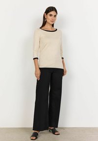 Beige fitted top with three-quarter sleeves, black trim at neckline and sleeve cuffs, paired with black wide-leg pants and black mules.