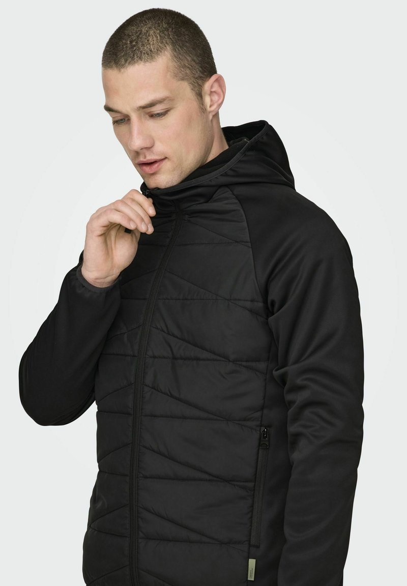 Veste à capuche noire avec fermeture éclair sur le devant, présentant une texture matelassée, un tissu lisse et un cordon de serrage à la capuche. Poche latérale incluse.
