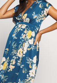 Raska kvinna i en blå blommönstrad klänning med V-ringning, ärmlös design, hand vilande på höften, och ett silverhalsband.