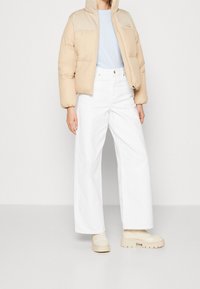 Béžová péřová bunda s kapucí a kontrastními rameny, spárovaná s bílými kalhotami s širokými nohavicemi a robustními krémovými botami.