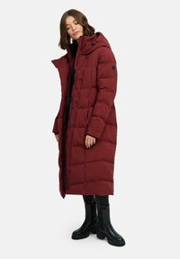 Lungo cappotto imbottito rosso con cappuccio, caratterizzato da un design trapuntato, bottoni a pressione e un'etichetta nera. Indossato sopra un abbigliamento nero e stivali.