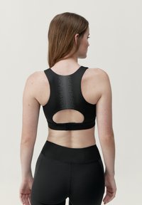 Svart sportbh med racerback-design, som har en urtagning och prickigt mönster ned längs ryggen. Tillverkad av elastiskt, slätt material.