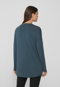 Langarmshirt in dunkeltürkis, aus weichem Stoff gefertigt. Mit einer lockeren Passform, rundem Ausschnitt und abgerundetem Saum, kombiniert mit schwarzen Leggings.