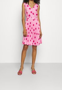 Robe plissée rose avec de grands pois, décolleté en V, sans manches et une ceinture assortie. Portée avec des sandales à talons roses.