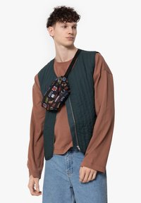 Quilted green vest with zip, worn over a brown long-sleeve shirt. Black patterned fanny pack attached to the vest. Blue shorts.