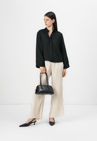 Black collared button-up shirt, cream pleated trousers, holding a small black handbag, complemented by black pointed-toe heels.
