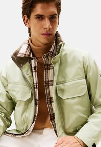 Chaqueta verde clara con dos bolsillos frontales, usada sobre una camisa de cuadros. El modelo tiene un cuello de tortuga marrón visible debajo de la camisa.