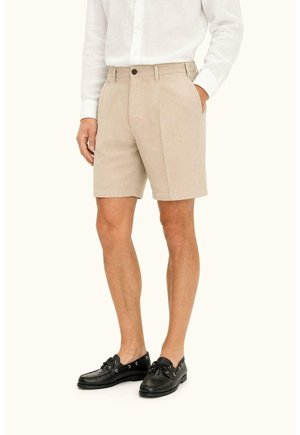 Man draagt beige shorts, een wit lang shirt en zwarte leren bootschoenen, staat met één hand in zijn broekzak.