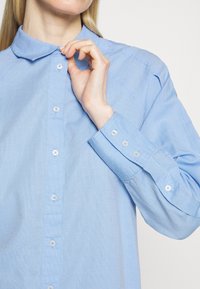 Personne en train de boutonner le col d'une chemise à manches longues texturée bleu clair avec un col classique devant un fond uni.