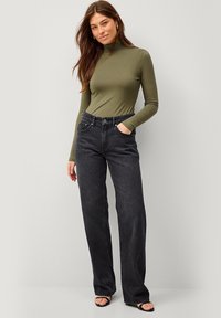 Olivgrünes Langarm-Turtleneck-Oberteil kombiniert mit hoch taillierten schwarzen Jeans aus Denim, die über Taschen und einen geraden Beinschnitt verfügen, vor einem neutralen Hintergrund.