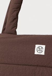 Hnědá tkaná taška s dvojitými uchy a malou bílou čtvercovou etiketou s černým kruhovým logem našitým na přední straně.