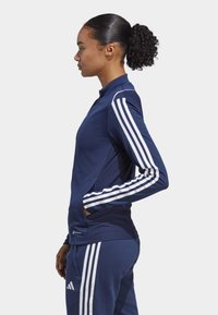Marinefarbener Sportjacke mit weißen Streifenakzenten, taillierter Schnitt, Reißverschlusskragen und strukturierten Seitenpanelen, kombiniert mit passenden Hosen.