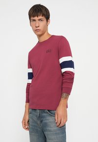 Maroon long-sleeve shirt with white and navy stripes on the sleeves, featuring a small black "GAP" logo on the chest. Cotton material.