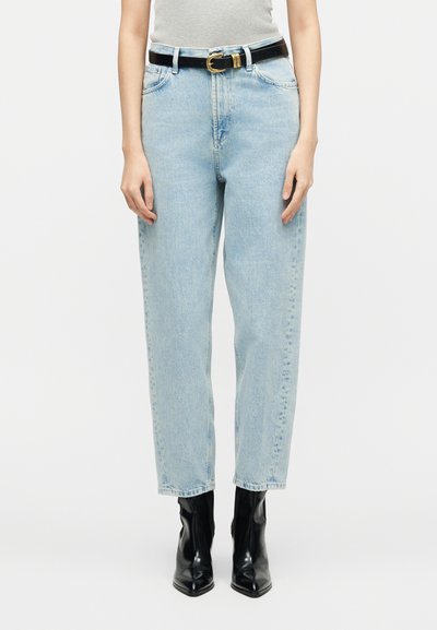 Άτομο που φορά ψηλόμεσα ανοιχτό μπλε τζιν, μαύρη δερμάτινη ζώνη και μαύρα μποτάκια με μυτερό μύτη, στέκεται μπροστά σε λευκό φόντο.