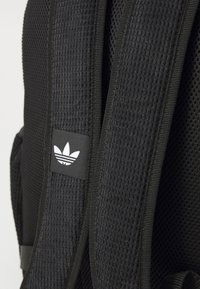 Rucsac negru cu un design din plasă texturată, bretele ajustabile și un mic logo Adidas alb pe un patch negru.