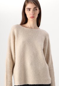 Šviesiai rudas megztinis su plačiu iškirpimu, ilgomis rankovėmis ir tekstūruotu, šiek tiek raukšlėtu paviršiumi. Minkšta medžiaga ir laisvas siluetas.
