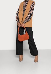 Tan sleeveless blazer over a floral black blouse, paired with wide-legged black pants and light brown ankle boots. A small orange suede handbag is held.